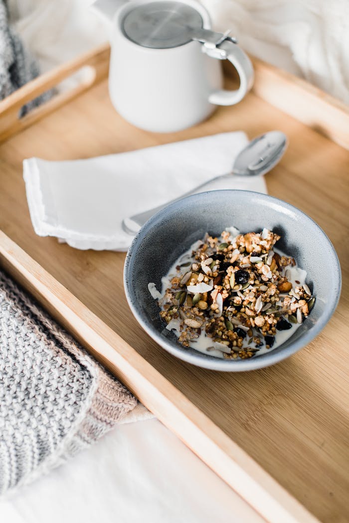From above of bowl with muesli with milk and small teapot placed on wooden tray placed on bed in morning time